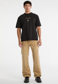 T-shirt di cotone nero con logo dorato "GUESS", abbinata a pantaloni cargo beige con tasche e scarpe nere. Design semplice e moderno.