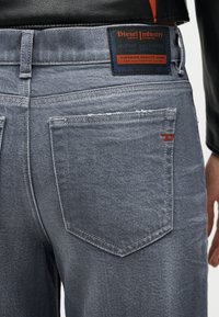 Πίσω μέρος ανοιχτόχρωμων τζιν denim με ορθογώνια ετικέτα Diesel Industry πάνω από την δεξιά τσέπη και κόκκινη ραφή στη χείλη της τσέπης.