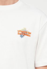 Λευκό πουκάμισο με μικρό γραφικό μπλε συστάδας σταφυλιών και πορτοκαλί κείμενο "Viñedos del Sol Dorado" στο στήθος.