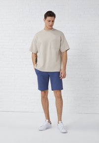 Beiges T-Shirt mit kurzen Ärmeln und einer Brusttasche, kombiniert mit blauen Shorts. Das Modell trägt weiße Sneakers. Der Hintergrund ist eine weiße Ziegelwand.