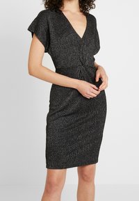 Svart klänning med silver vertikala ränder, korta svängande ärmar, V-ringning, rynkad midjedetalj och figurnära silhuett. Slät textur.