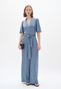 Hellblauer Jumpsuit mit kurzen Ärmeln und Wickeldesign, gebundenem Gürtel, weitem Bein, glatter Textur, kombiniert mit einer schwarzen Clutch und Sandalen.