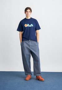 T-shirt en coton bleu marine avec un logo "Carhartt" coloré, associé à un pantalon en denim ample et des chaussures à lacets orange avec des semelles texturées.