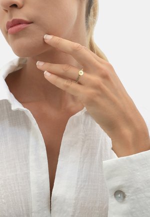 Anello d'oro con un disco piano rotondo che presenta la lettera "S," indossato su una mano vicino al viso. Il modello è vestito con una camicia di lino bianca.