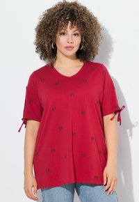 T-shirt rouge en V en tissu doux avec des motifs de nœuds noirs, doté de manches roulées avec des liens. Porté avec un jean bleu.