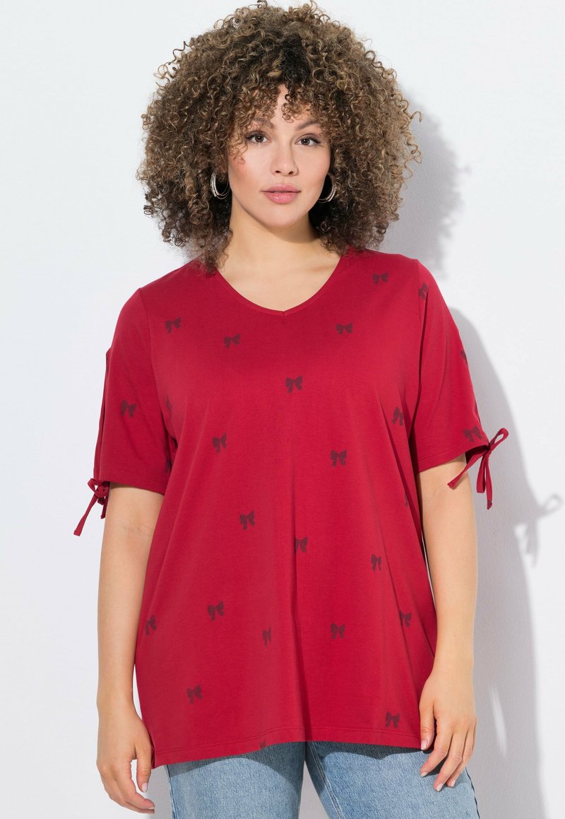 T-shirt rouge en V en tissu doux avec des motifs de nœuds noirs, doté de manches roulées avec des liens. Porté avec un jean bleu.