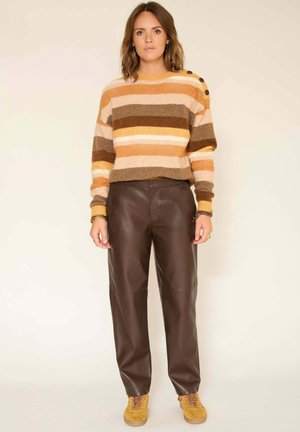 Pull en tricot rayé orange, marron et crème avec des boutons aux épaules, associé à un pantalon en cuir marron et des baskets en daim jaunes.