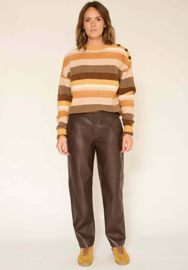 Pull en tricot rayé orange, marron et crème avec des boutons aux épaules, associé à un pantalon en cuir marron et des baskets en daim jaunes.