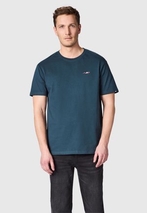 Man draagt een donker teal kortärmelig T-shirt met een klein rood-wit logo op de linker borst, gecombineerd met zwarte jeans.