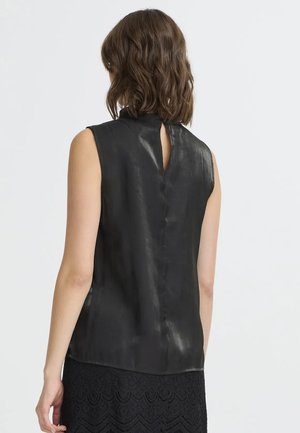 Frau mit schulterlangem welligem Haar trägt ärmelloses schwarzes Satin-Oberteil mit Schlüsselloch am Rücken und schwarzen Spitzenrock, mit dem Rücken zur Kamera.