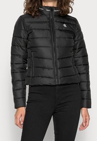 Svart, vadderad jacka med ståkrage, dragkedjestängning och logodetalj. Har horisontella sömmar och en figursydd siluett.