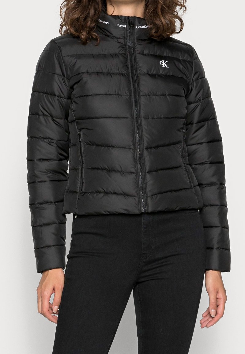 Svart, vadderad jacka med ståkrage, dragkedjestängning och logodetalj. Har horisontella sömmar och en figursydd siluett.