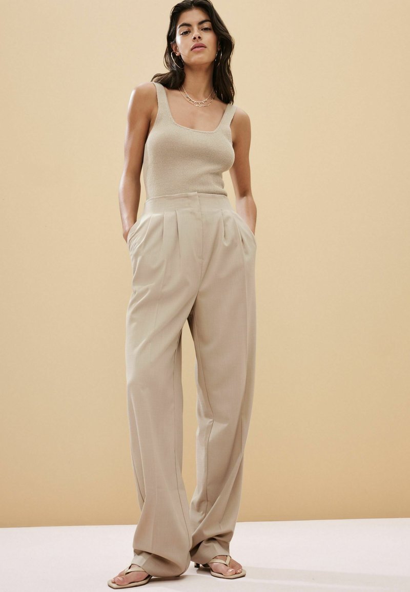 Beige ribbet tank top og højtsiddende plisserede bukser i en lys beige farve, parret med flade beige sandaler. Simpelt design og tekstur.