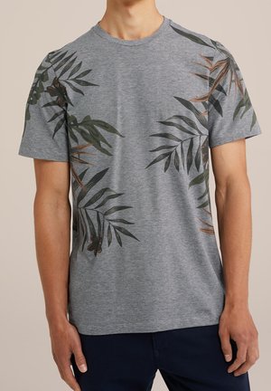 Mann trägt graues T-Shirt mit kurzen Ärmeln und dunkelgrünen und braunen tropischen Blättern.
