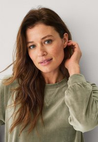 Felpa verde oliva, morbida, con maniche a sbuffo e scollo rotondo, indossata da una donna con capelli lunghi e mossi, su uno sfondo neutro.
