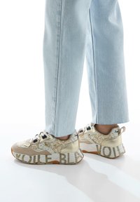 Chunky sneakers in gold and beige, featuring a textured upper, black accents, and bold "VOILE" lettering on the sole. Paired with light blue jeans.