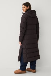 Lange, donkerbruine pufferjas met capuchon, voorzien van horizontale stiksels en zijritsen. Draagt over losse, donkere jeans en tan kleurige laarzen.