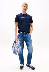 Tamsiai mėlynas Tommy Jeans marškinėlis su logotipu, mėlyni džinsai, laikantis languotą šviesiai mėlyną flanelinį marškinį ir dėvintis tamsiai mėlynas sportbačius su baltais akcentais.