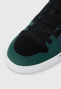 Sneaker met een zwarte canvas bovenkant, groene leren accenten, geperforeerde neuspartij en witte rubberen zool met een gestructureerde grip.