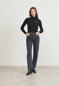 Maglione zip-up nero a coste con un piccolo logo, abbinato a jeans grigi a vita alta e stivaletti neri. Sfondo semplice.