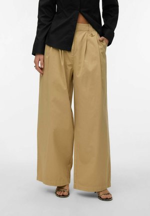 Beige bukser med vide ben og frontplissering, båret med en sort skjorte med knapper og åbne sandaler med hæl, hænderne i lommerne.