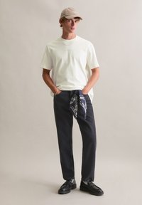 Biała bawełniana koszulka, czarne spodnie z niebieską chustką w wzory zawiązaną w pasie oraz czarne mokasyny. Neutralne tło.