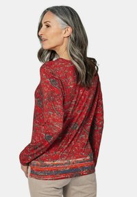Rotes Langarmshirt mit Paisleymuster, weichem Stoff und abgerundetem Saum. Auf der Rückseite ist ein mehrlagiges Design mit kontrastierenden Streifen zu sehen.