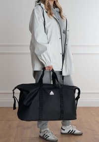 Borsone nero con due manici robusti, caratterizzato da un'etichetta con logo, realizzato in materiale liscio, portato da una persona in abbigliamento grigio.
