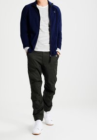 Giacca zip-up blu navy sopra una maglietta a righe grigie e bianche, abbinata a pantaloni larghi verde scuro e scarpe da ginnastica bianche.