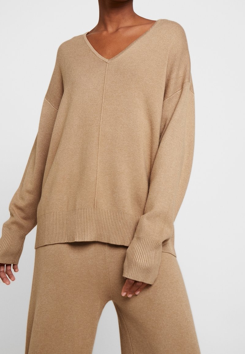 Personne portant un pull en maille beige ample à col en V avec poignets et ourlet côtelés, associé à un pantalon en maille assorti.