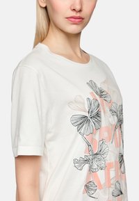 White cotton t-shirt featuring black floral embroidery and pink graphic text. Short sleeves, crew neck, smooth texture, and relaxed fit.