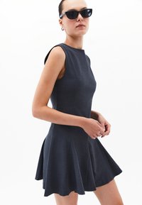Vestido sin mangas de color gris oscuro con un torso ajustado y una falda acampanada. Hecho de una tela texturizada, presenta un escote limpio y aberturas laterales.