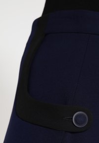 Vêtement bleu marine avec une ceinture noire, détail de bouton rond et un tissu lisse et texturé. La forme est cintrée.