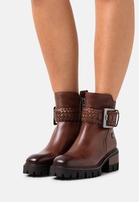 Hnědé kožené kotníkové boty s pleteným pásem a stříbrnou sponou, texturovaný povrch, robustní černá podrážka a zaoblený design špičky.