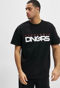 Melns kokvilnas T-krekls ar izteiksmīgu baltu un sarkanu grafiku ar tekstu "DANGEROUS DNGRS", kas novietots centrā uz priekša. Īsās piedurknes.