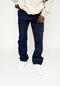 Pantalones cargo de color azul marino hechos de una tela resistente, con múltiples bolsillos, que presentan un corte relajado y se estrechan en los tobillos. Combinados con zapatillas blancas.