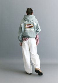 Miętowa bluza z kapturem z białym napisem, w oversize'owym fasonie, nałożona na czerwono-biały pasiasty longsleeve. Białe szerokie spodnie i brązowe buty.