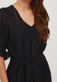 Černé šaty s výstřihem do V, texturovanou tkaninou s vodorovnými pruhy, krátkými nabíranými rukávy a zavazovacími pásky v pase.
