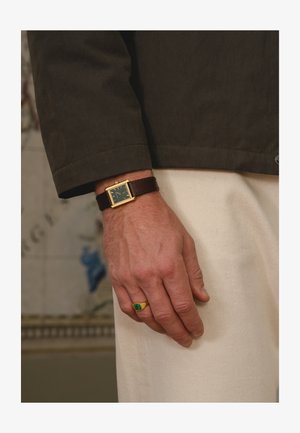 Męska ręka nosząca złoty prostokątny zegarek z brązowym skórzanym paskiem oraz złoty pierścionek z zielonym kamieniem, spoczywająca na kremowych spodniach.