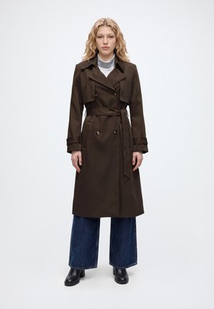 Un mannequin se tient devant un fond uni, vêtu d’un trench-coat marron foncé ceinturé, d’un jean à jambes larges et de bottes noires.