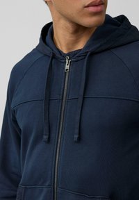 Felpa con zip di colore blu navy, dotata di cappuccio regolabile con cordino, texture morbida e dettagli di cucitura a contrasto lungo il petto e le spalle.