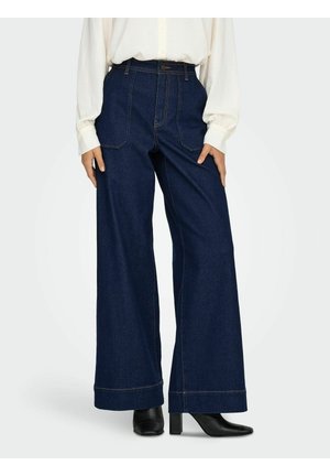 Pantalons en denim beige à jambes larges, couleur bleu foncé, avec des coutures jaunes contrastantes, taille haute et ourlets repliés, associés à un chemisier blanc à manches longues.