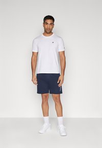 Camiseta blanca de manga corta con un logo en el pecho, combinada con pantalones cortos azul marino con detalles laterales. Usada con zapatillas blancas y calcetines.
