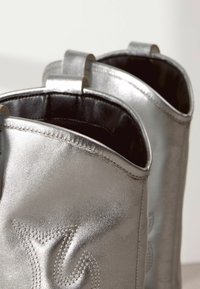 Sidabriniai metaliniai kulkšninių bateliai su lygaus paviršiaus, išsiskiriantys kontrastuojančia siuvimo linija ir traukimo kilpomis gale.