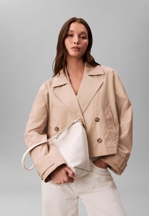 Kvinne iført beige kort dobbelspent jakke og hvite bukser, holder en stor hvit håndveske mot en ensfarget grå bakgrunn.