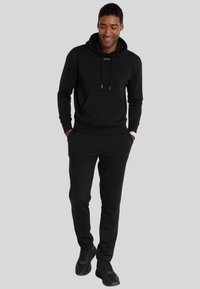 Felpa nera con tasca frontale, coulisse regolabile e dettaglio del logo. Abbinata a pantaloni jogger neri coordinati e sneakers nere.