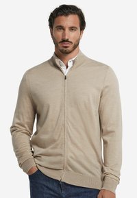 Beige zip-up cardigan med hög krage, tillverkad av mjukt tyg, med en slät yta och ribbade ärmmuddar, bärs över en vit skjorta.