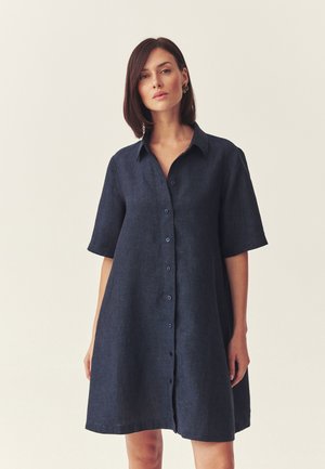 Kobieta ubrana w luźną, krótkorękawową, granatową sukienkę zapinaną na guziki z kołnierzykiem, stojąca na tle jednolitego jasnego tła.