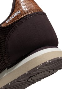 Gros plan sur le talon de la basket marron montrant le cuir texturé, le tissu noir, la broderie du logo blanc, et une semelle superposée beige et marron foncé.