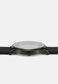 Runder schwarzer Uhr mit einem strukturierten silbernen Zifferblatt, klarem Kristall und einem mattschwarzen Silikonarmband. Verfügt über eine einzelne Krone an der Seite.